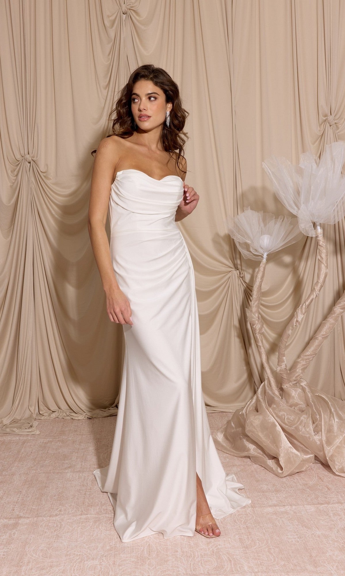 A woman wears the Long Prom Dress: Eureka EK178, standing barefoot on a light pink floor with beige draped curtains and a decorative tree in the background.